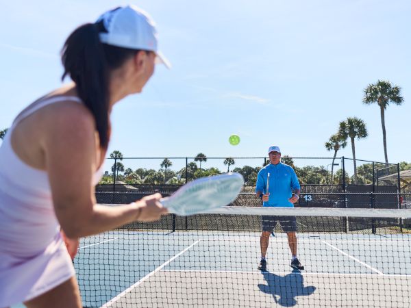 A woman and a man playing pickleball on a sunny day, with a net and palm trees visible in the background.
