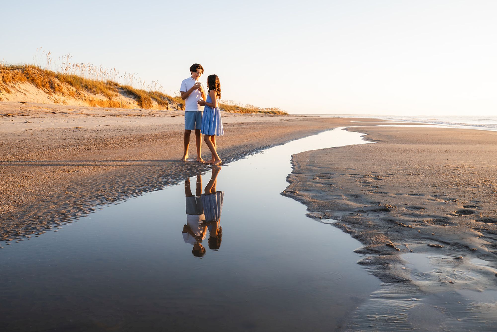 A couple stands on a beach by a shallow water stream, with their reflection visible, under a clear sky at sunset.