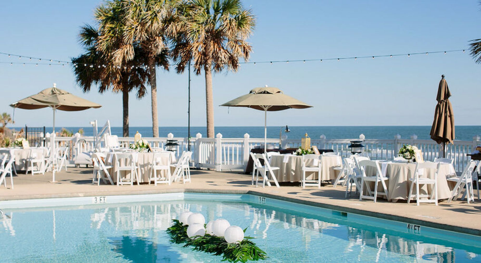 An outdoor event setup by the sea with tables, chairs, umbrellas, and palm trees surrounding a pool decorated with greenery and white spheres.