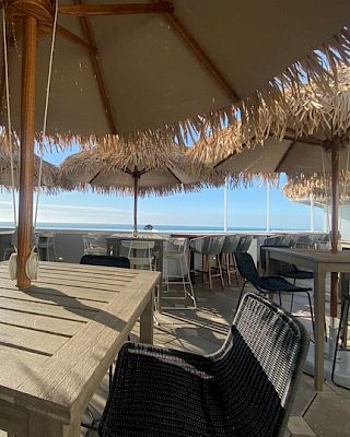 An outdoor dining area with wooden tables, chairs, and straw umbrellas overlooks a scenic view in the background.