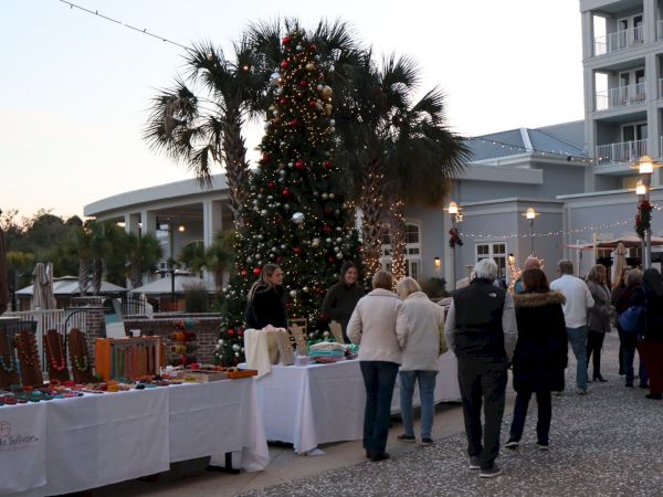 A group of people is gathered near a decorated Christmas tree with tables displaying items, set outdoors with buildings in the background.
