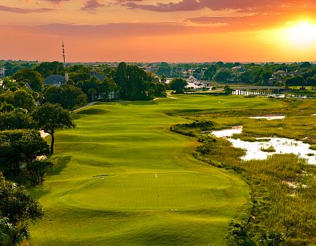 A scenic sunset over a lush golf course, with greens, fairways, trees lining the left, a waterlogged marsh on the right, and calm houses beyond.