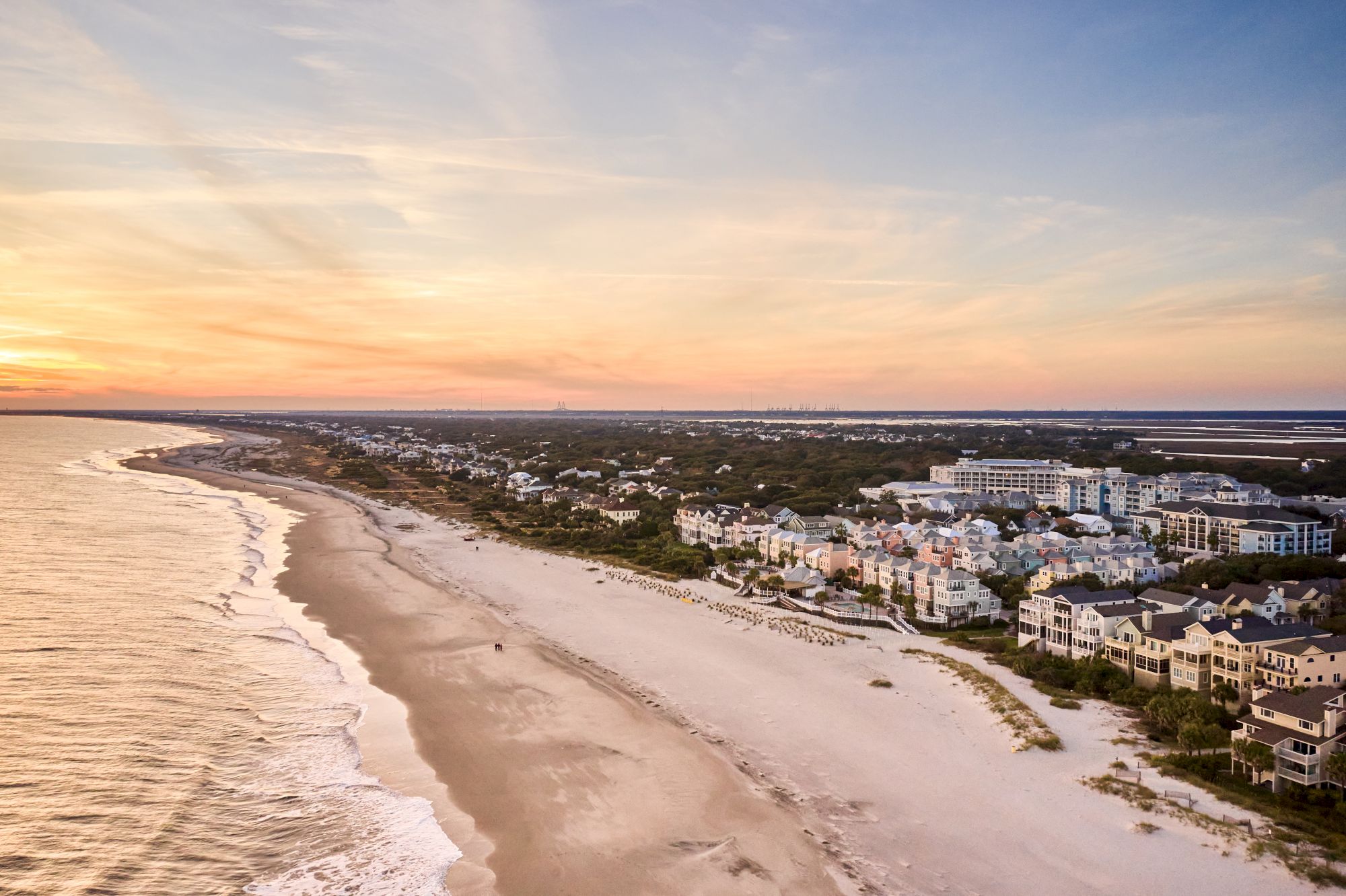 The image shows a coastal town with a sandy beach, ocean waves, and a sunset sky, alongside houses and lush greenery.