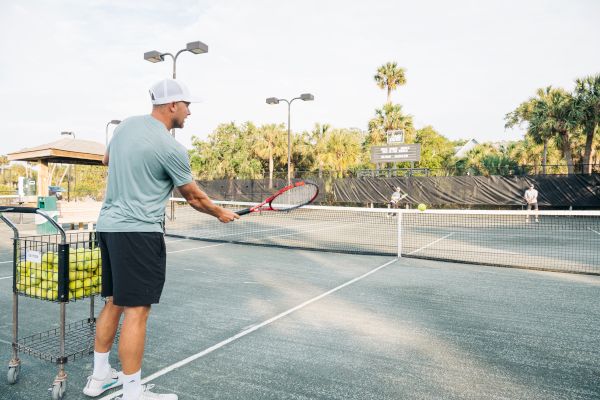 A person on a tennis court holds a racquet near a cart filled with tennis balls, with another person at the far end of the court.