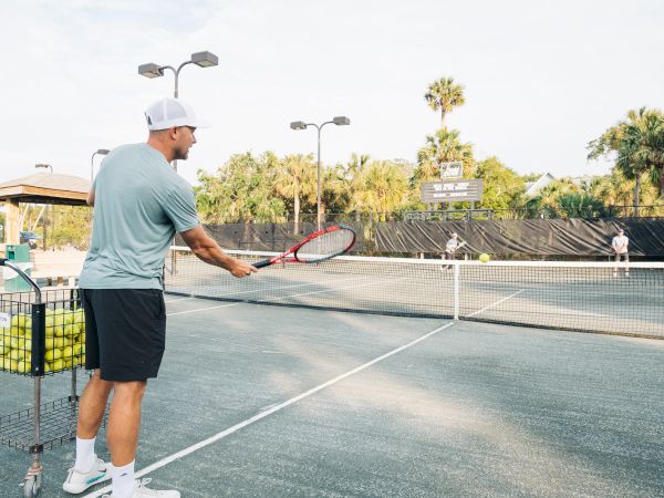 A person on a tennis court holds a racquet near a cart filled with tennis balls, with another person at the far end of the court.