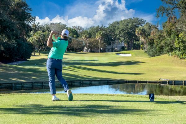 A golfer in a teal shirt is teeing off on a sunny golf course, with trees and a sand trap in the background.