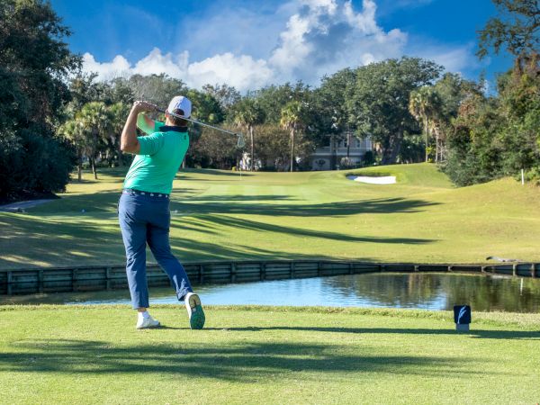 A golfer in a teal shirt is teeing off on a sunny golf course, with trees and a sand trap in the background.