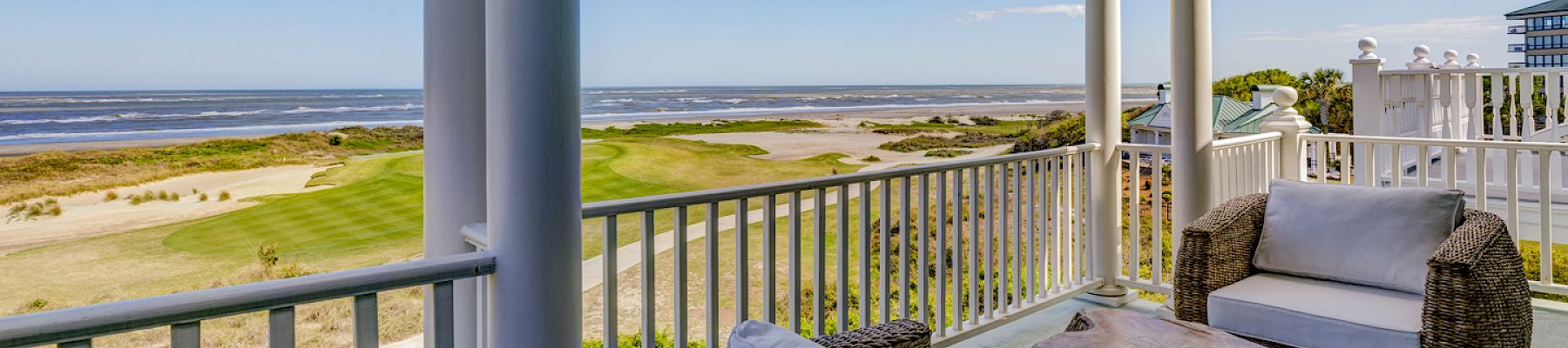 A porch with wicker chairs overlooks a golf course, sandy area, and ocean under a clear blue sky, offering a serene coastal view.