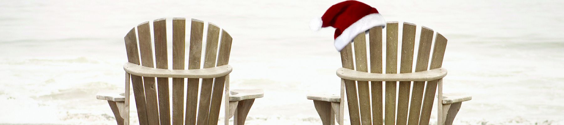Two wooden Adirondack chairs facing the ocean, one with a Santa hat placed on it, suggesting a festive beach scene.