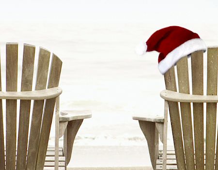 Two wooden Adirondack chairs facing the ocean, one with a Santa hat placed on it, suggesting a festive beach scene.