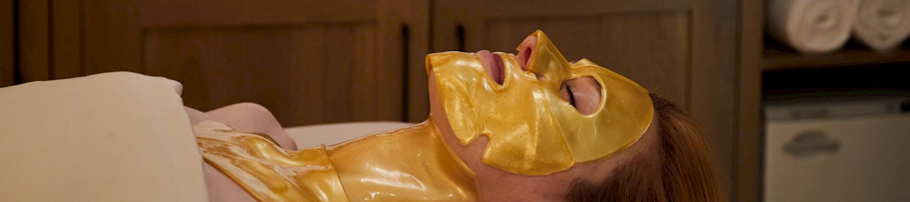 A person is relaxing on a spa bed with a gold facial mask, covered by a sheet, in a serene spa setting with towels and products nearby.