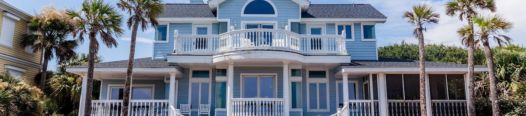 A blue coastal house with white trim, surrounded by palm trees, features multiple balconies and large windows under a clear sky.