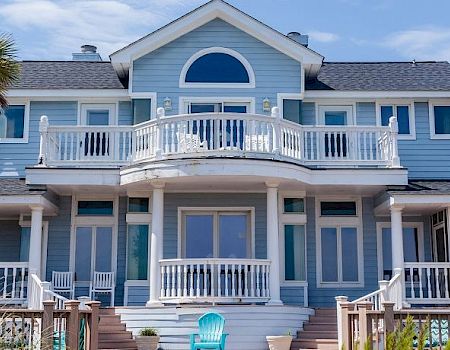 A blue coastal house with white trim, surrounded by palm trees, features multiple balconies and large windows under a clear sky.