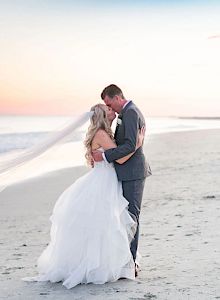 A bride and groom embrace on a beach at sunset, with the bride's veil flowing in the wind.