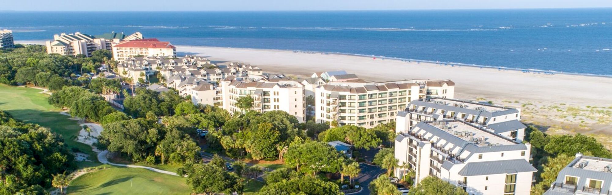 A coastal view with beachfront buildings, lush greenery, and a sandy beach extending to a calm blue sea under a clear sky.