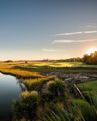 A serene landscape featuring a river, marshland, and a house with the sun setting in the background.