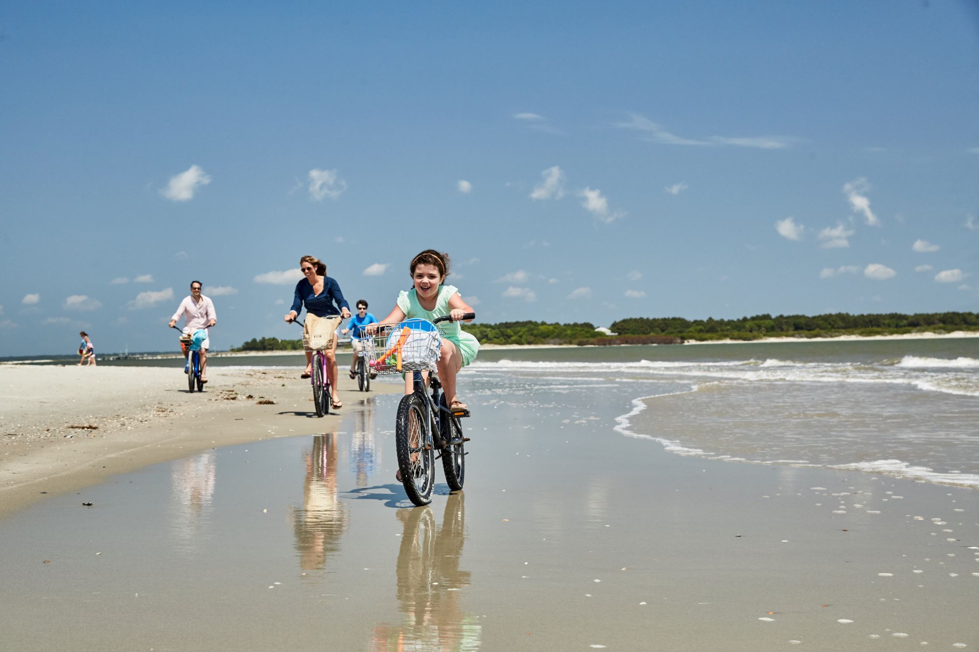 People are riding bicycles along a sandy beach near the water's edge under a clear blue sky with scattered clouds.
