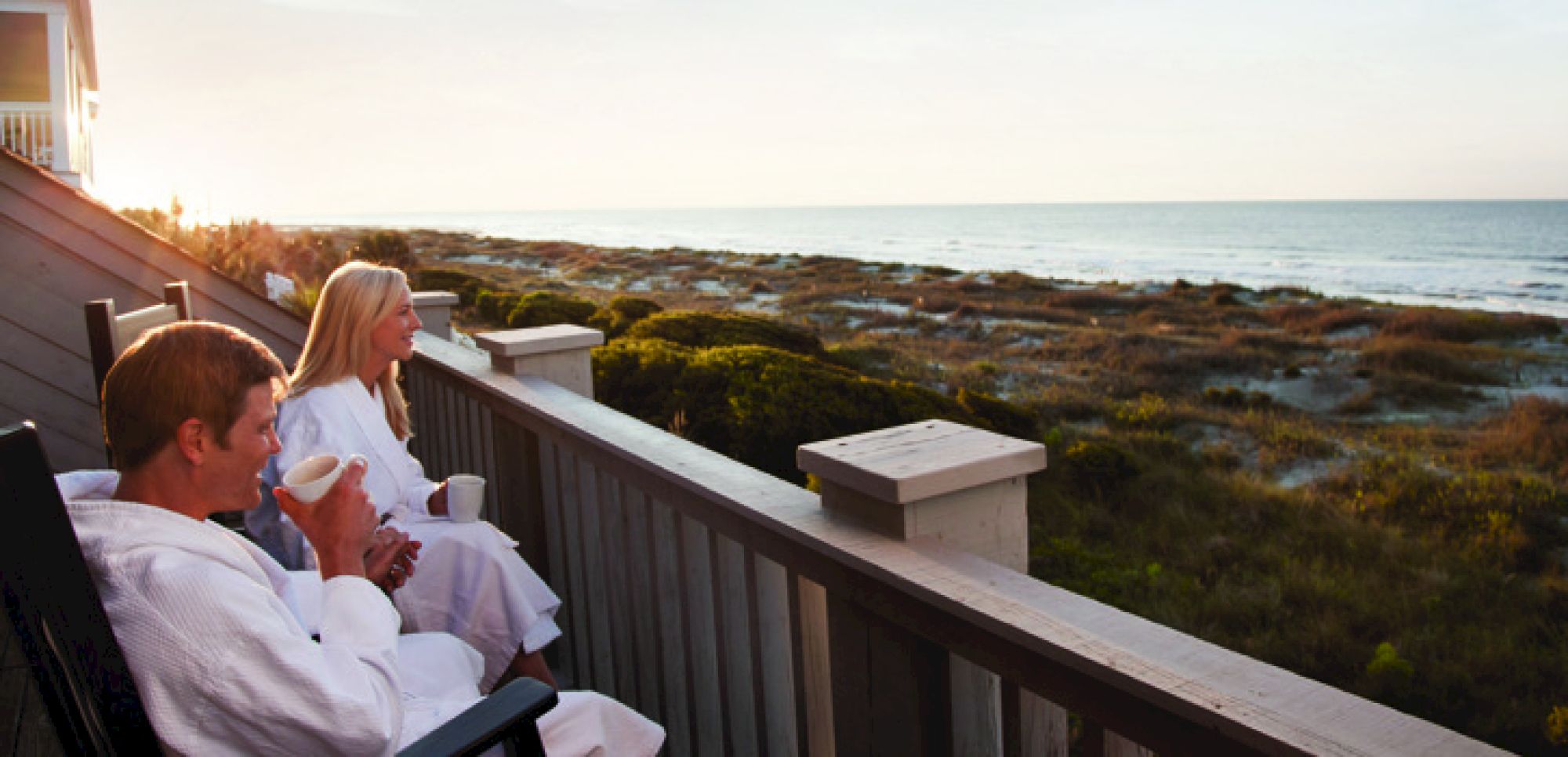 Two people in robes are sitting on a balcony, enjoying a drink while looking at the ocean view in the distance.