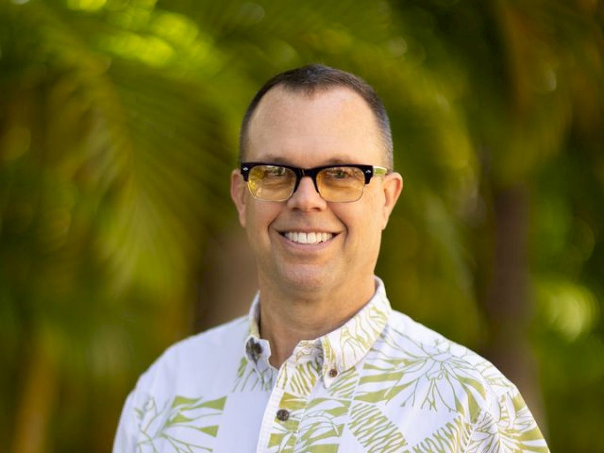 A person wearing glasses and a short-sleeve shirt with a leaf pattern stands smiling outdoors with a blurred green background.