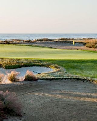 A scenic golf course with a sand trap in the foreground, lush green fairway, and ocean in the background under a clear sky.