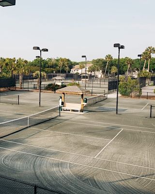 The image shows a deserted tennis court complex with multiple courts, surrounded by trees and lights, under a clear sky.