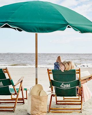 A beach scene with two chairs under a green umbrella. One chair is occupied, with a beach bag and hat nearby by the sea.