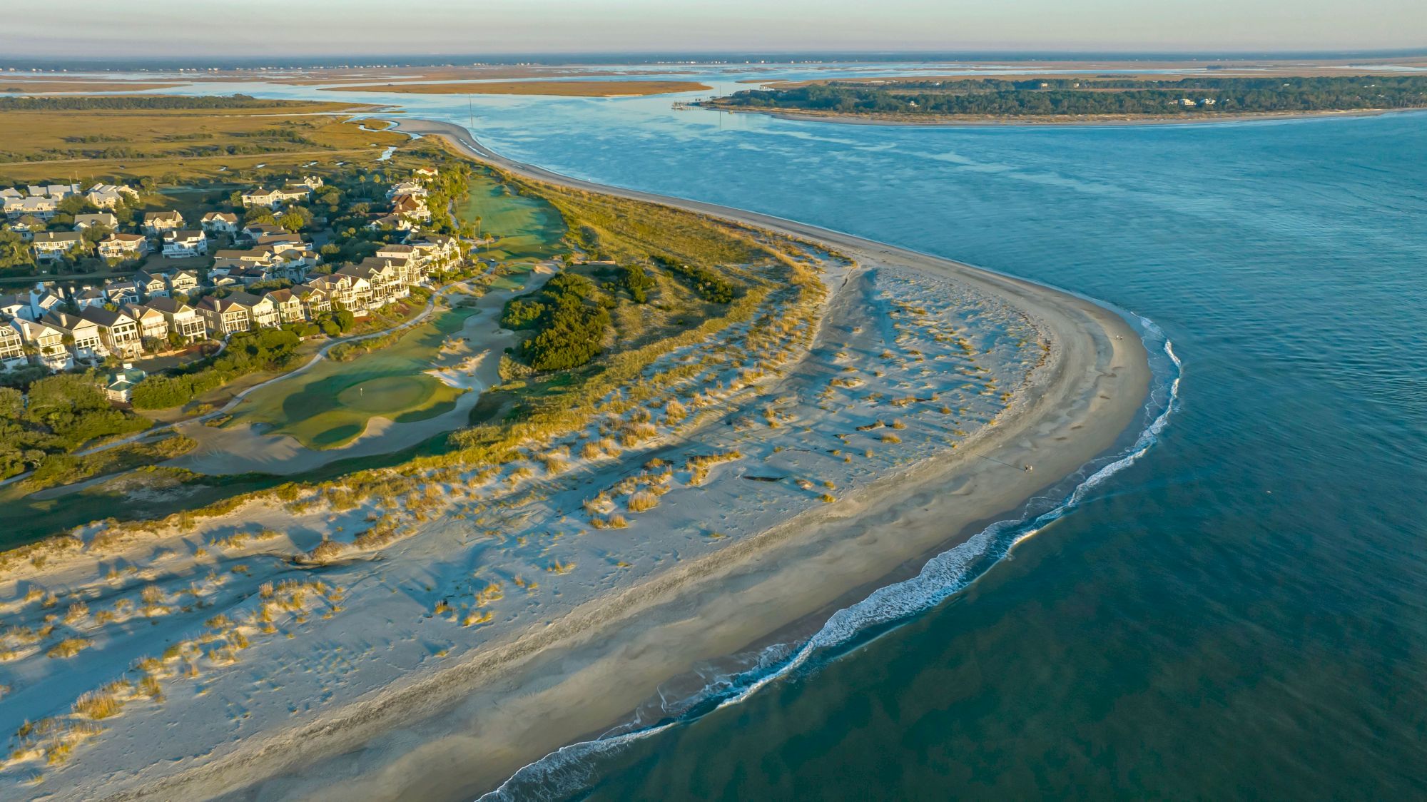 Aerial view of a coastal area with sandy beaches, gentle waves, and a residential neighborhood adjacent to a golf course and vegetation.