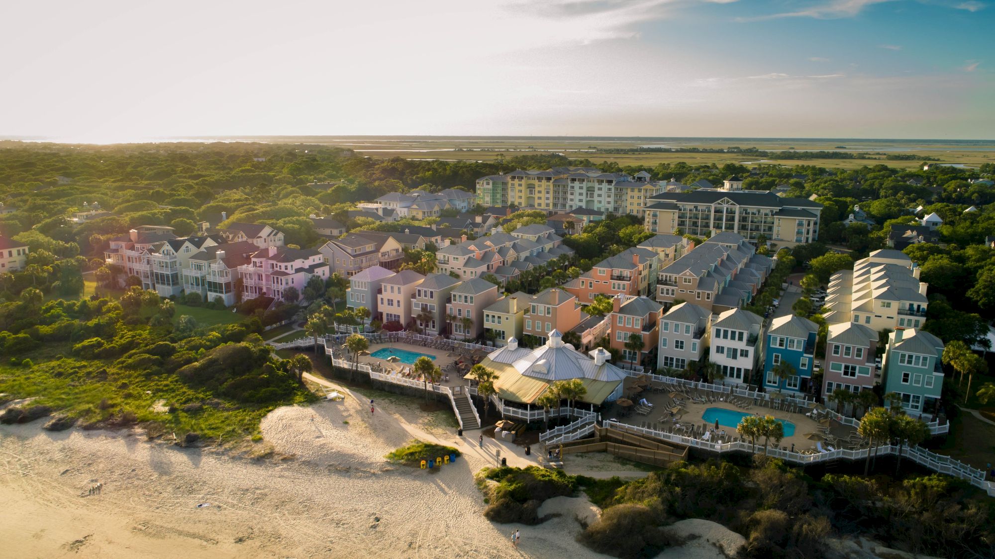Aerial view of a coastal resort town with colorful houses, two outdoor pools, and beach access, surrounded by lush greenery and ocean views.