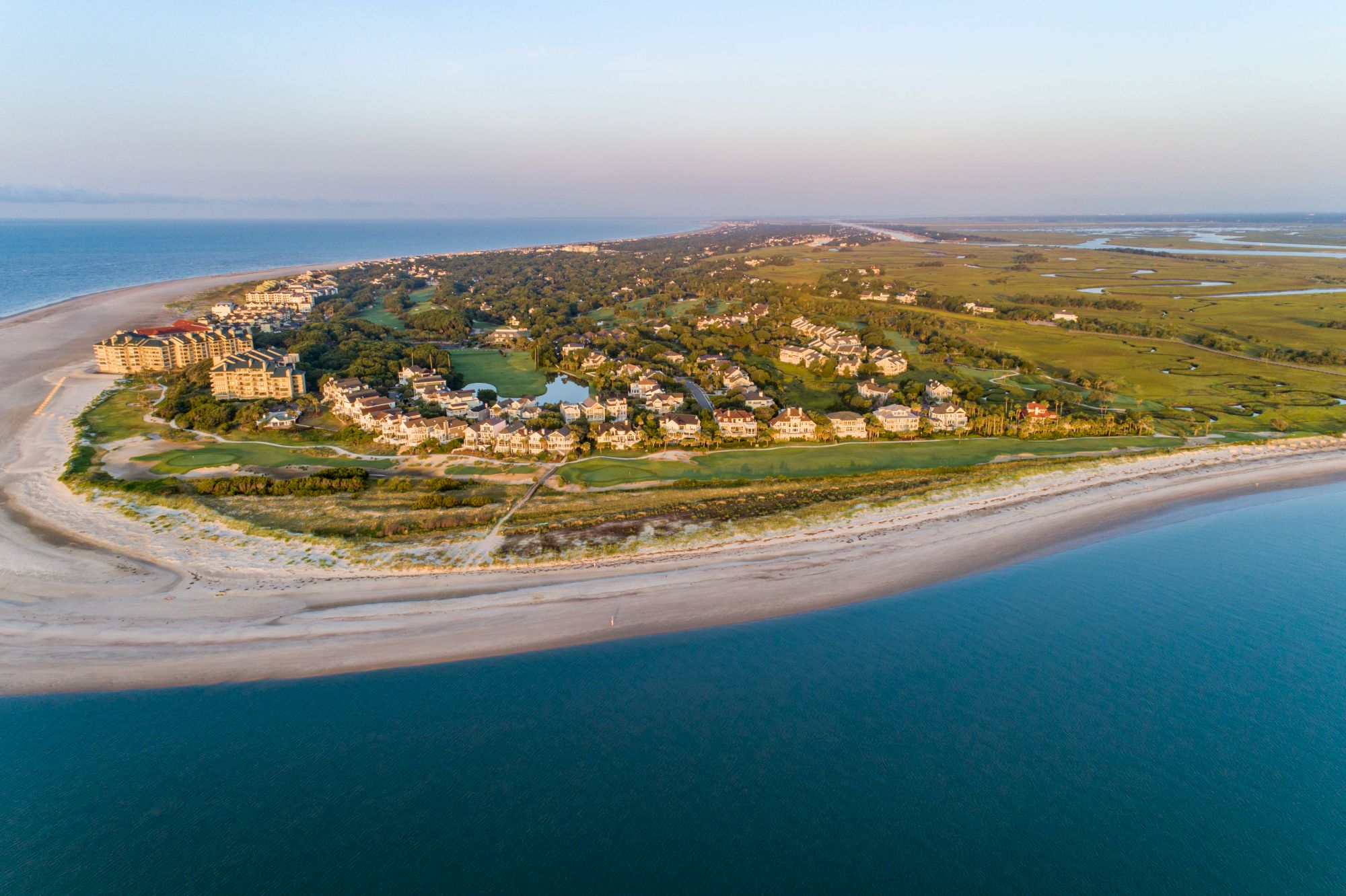 Aerial view of a coastal area with sandy beaches, blue ocean, residential buildings, green landscapes, and a golf course at sunset.