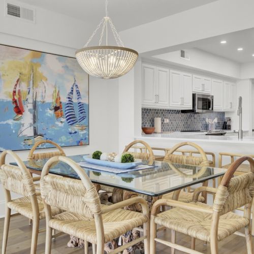 A modern dining area with a glass table, wicker chairs, a large nautical-themed painting, and a kitchen in the background with white cabinets.