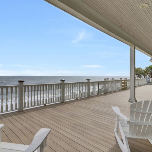 A beachfront deck with Adirondack chairs overlooks the ocean, framed by a railing and lined with palm trees under a clear blue sky.