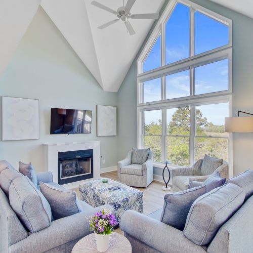 A modern living room with light blue sofas, a fireplace, large windows, and a vaulted ceiling.