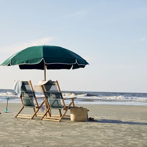 Two beach chairs and an umbrella face the ocean on a sandy beach, with a hat, towel, toy sailboat, and beach bag beside them.