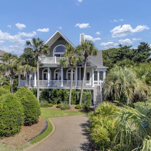 A picturesque house with palm trees, a lush garden, and a curved driveway under a blue sky with clouds.