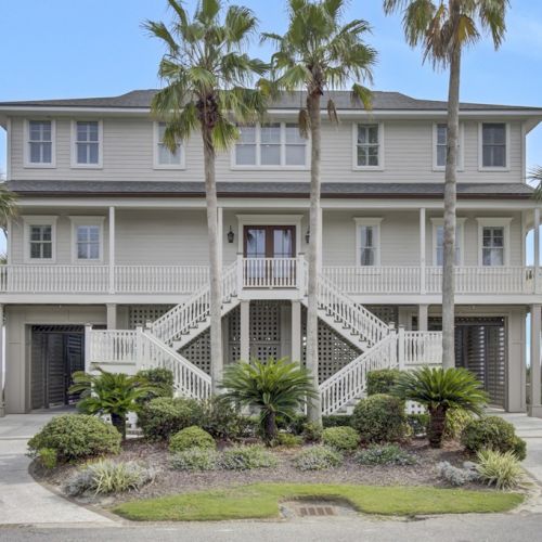 A large, two-story house with palm trees, featuring double staircases, a covered porch, and tropical landscaping in the front yard.