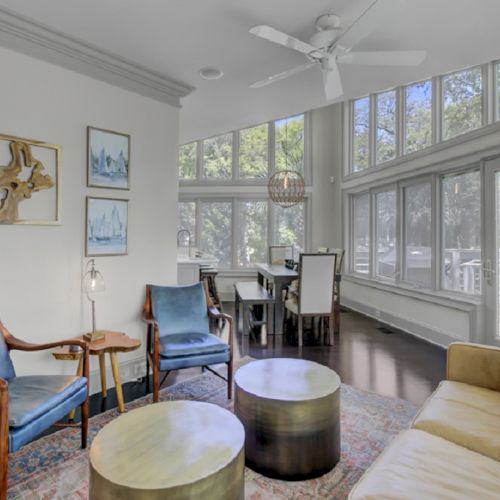 A stylish living room with modern furniture, large windows, blue chairs, and a dining area with a view, creating a bright, airy space.