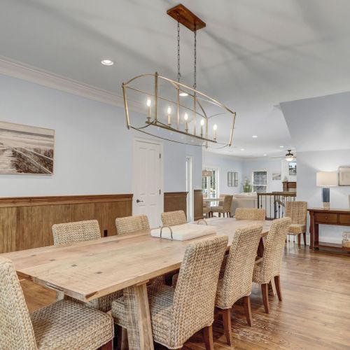 A spacious dining area with a large wooden table, woven chairs, and a modern chandelier. A landscape photo hangs on the wall.