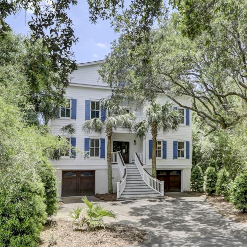 A two-story white house with blue shutters, surrounded by trees and greenery, featuring stairs leading to the entrance.