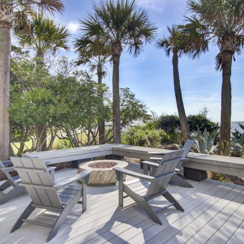 A coastal deck with gray chairs and a fire pit, surrounded by palm trees and greenery, overlooks the ocean under a clear blue sky.