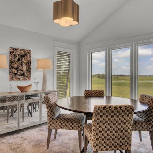 A dining area with a round table, patterned chairs, sideboard with lamps, artwork, large windows, and grassy field view.