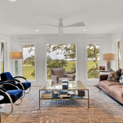 A modern living room with large windows, blue chairs, a brown sofa, artwork, and a glass coffee table, overlooking a scenic view outdoors.