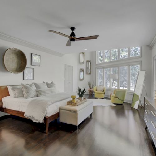 A modern bedroom with a bed, seating area, large windows, ceiling fan, and wall art, featuring neutral tones and minimalist decor.