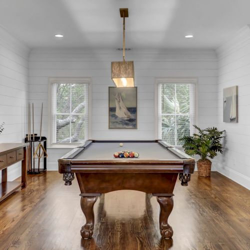The image shows a room with a pool table, wall decor of a fish, plants, paintings, and wooden furniture, featuring a windowed backdrop.