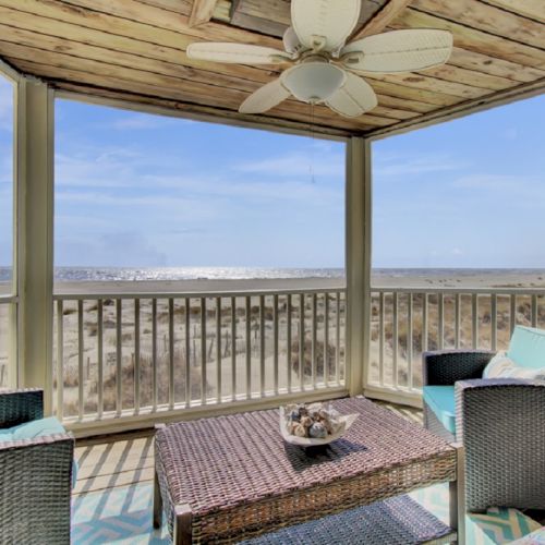 A seaside porch with wicker furniture, cushions, a ceiling fan, and a view of the ocean under a clear blue sky.