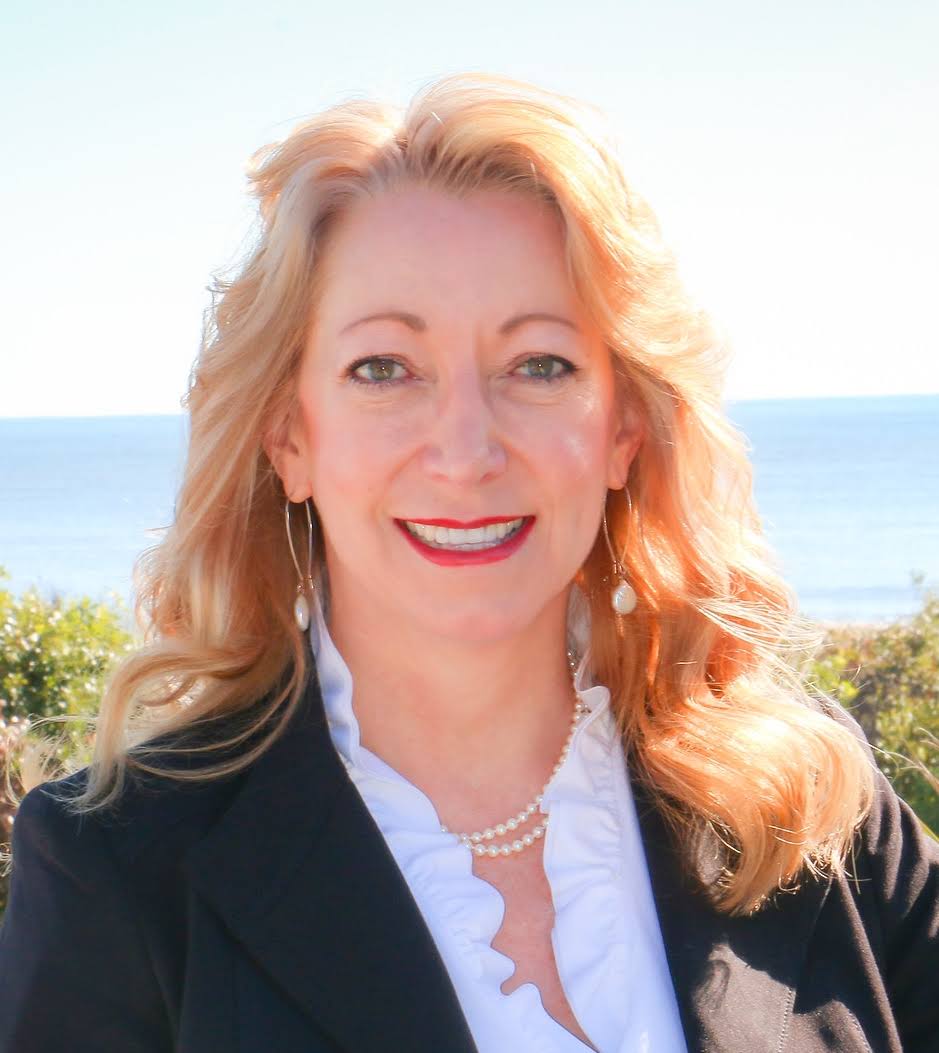 A woman with blonde hair smiles in a professional outfit, with a scenic ocean background.
