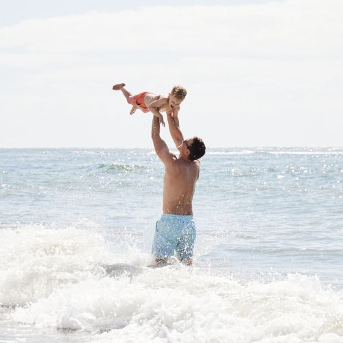 A person lifts a child above the waves in the ocean, creating a joyful scene under a cloudy sky at the beach.