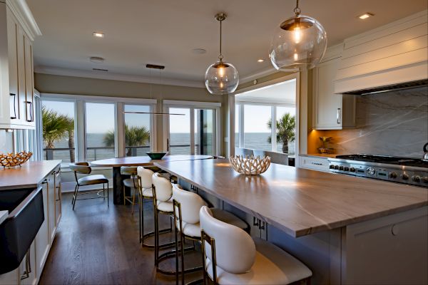 A modern kitchen with island seating, pendant lights, and ocean view through large windows, featuring light wood surfaces and white cabinetry.