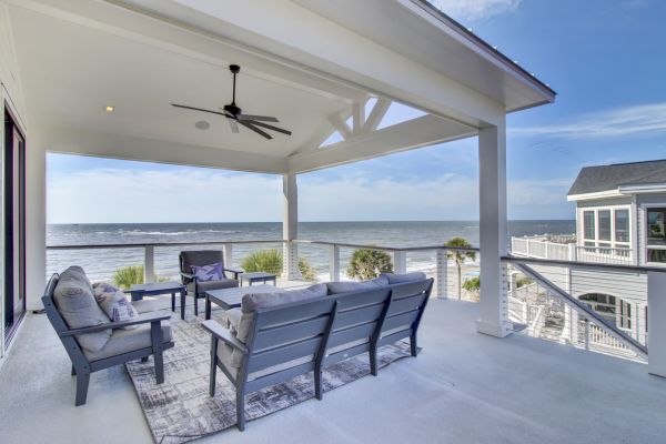 A covered deck with outdoor seating overlooks the ocean, featuring a ceiling fan and a view of nearby homes under a clear blue sky.