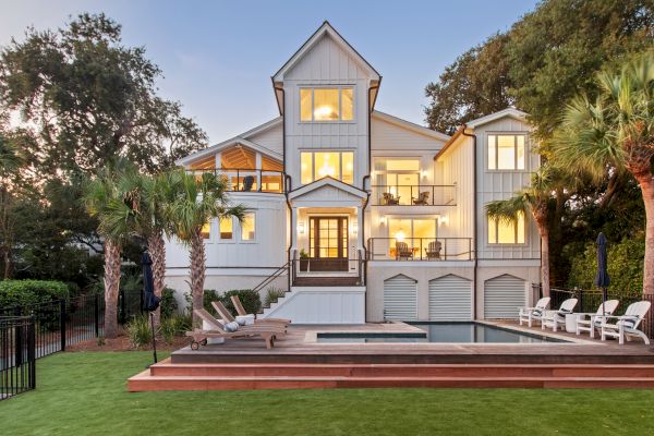 A modern, multi-story house features large windows, a pool, deck chairs, and is surrounded by trees and a well-kept lawn.