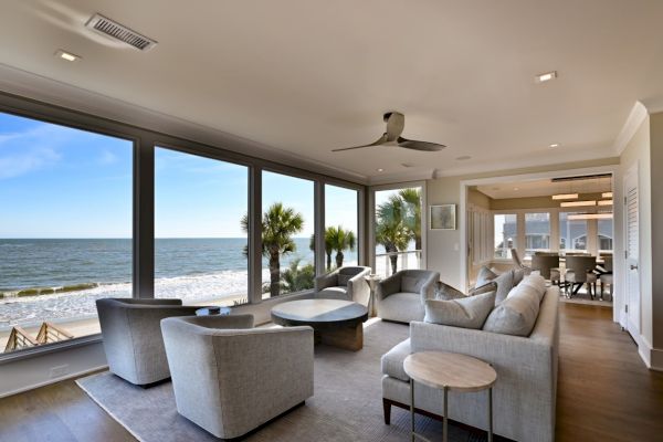 A modern living room with large windows offers a stunning ocean view, featuring gray sofas, armchairs, ceiling fan, and a dining area.
